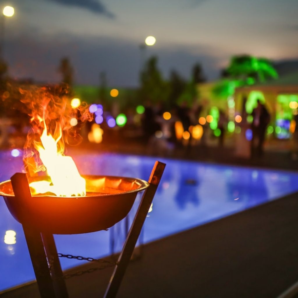 A close up of a fire feature with a pool and party goers in the background at night.